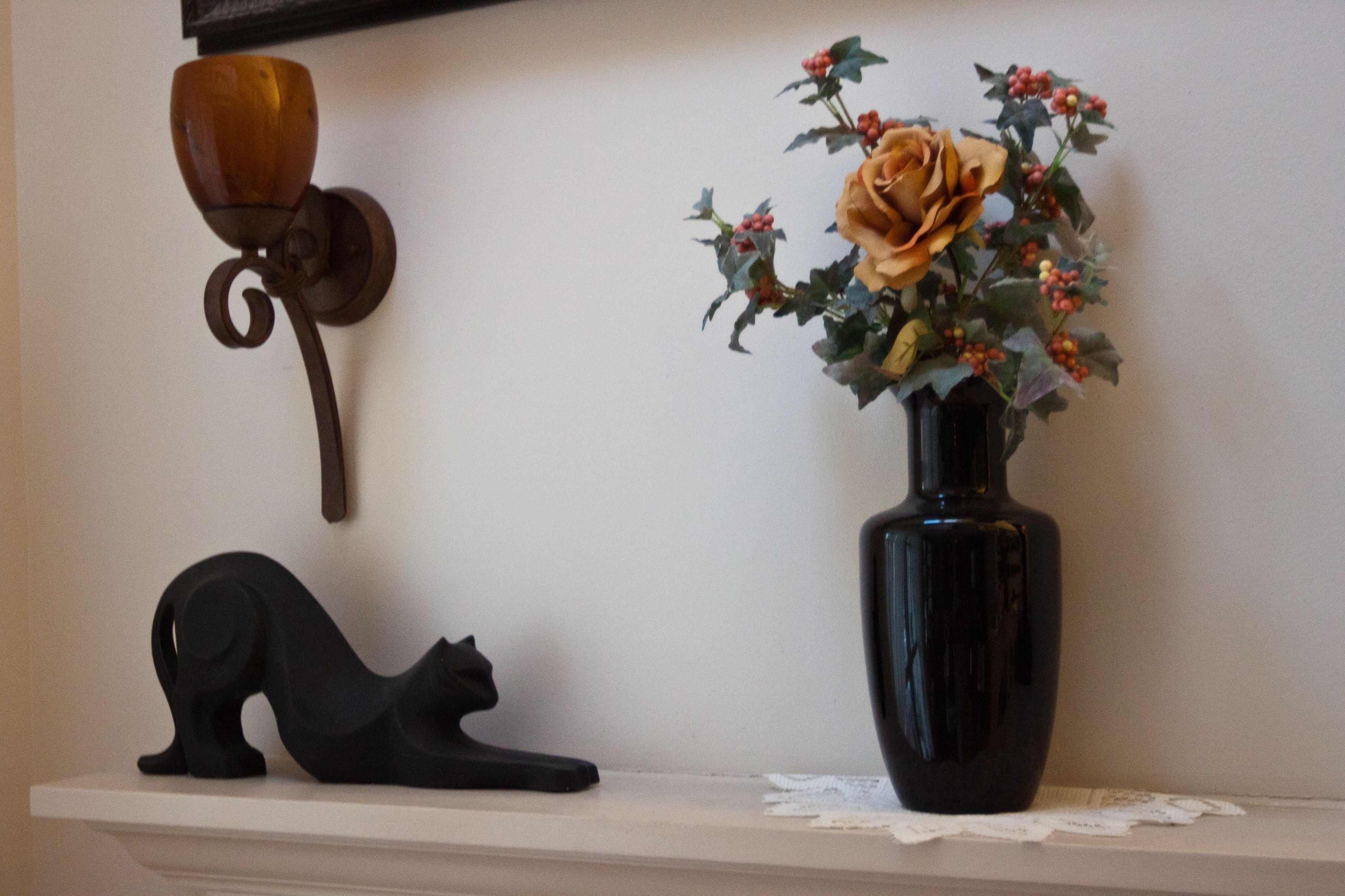 A black cat sculpture on a shelf, a vase with a rose, and a wall-mounted light fixture.