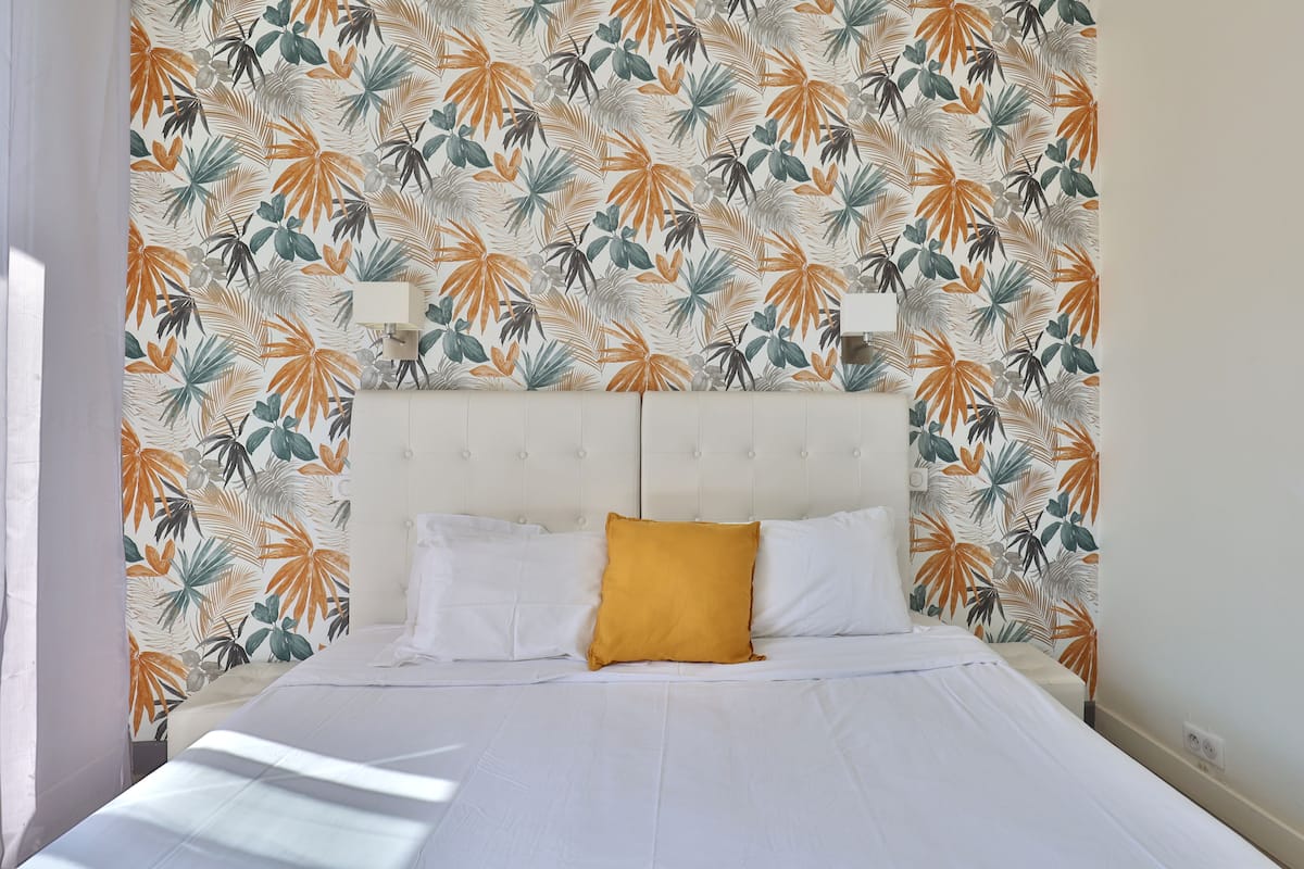 A neatly made bed with white linens and a mustard-colored pillow, set against a wall with a tropical leaf pattern.