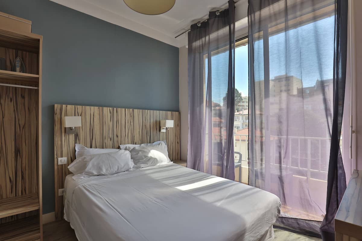 A bedroom with a bed, wooden headboard, and a view of buildings through sheer curtains.