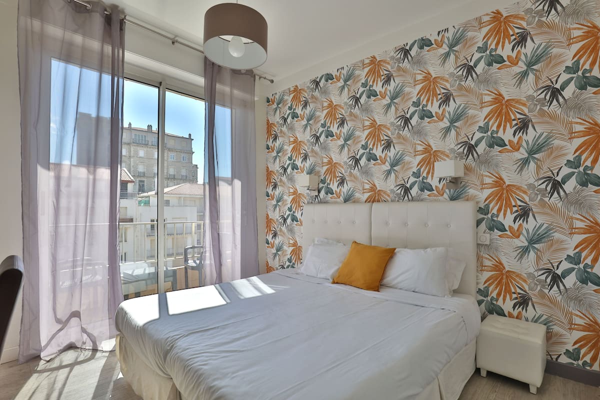 A bedroom with a large bed, a headboard, a bedside table, a window with curtains, and a wall with a tropical leaf pattern.