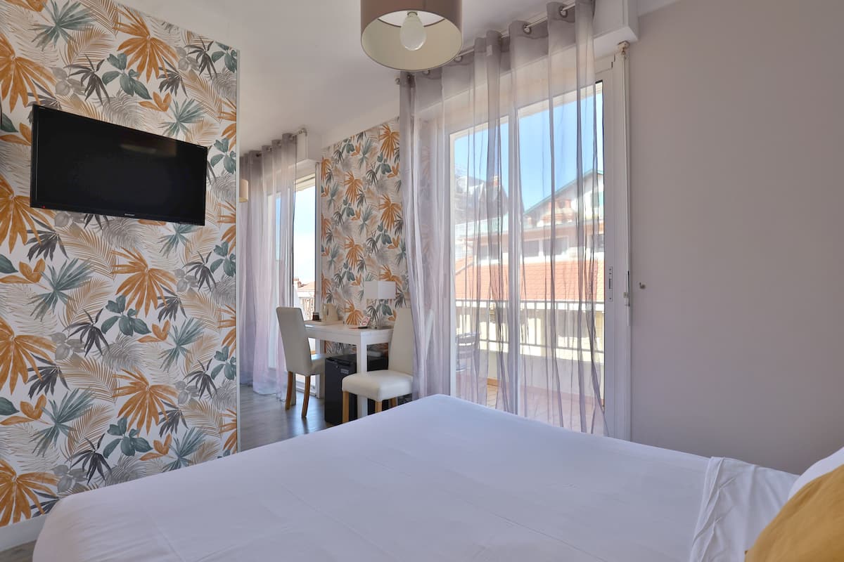 A hotel room with a bed, a desk with a chair, a television, and a balcony with curtains.