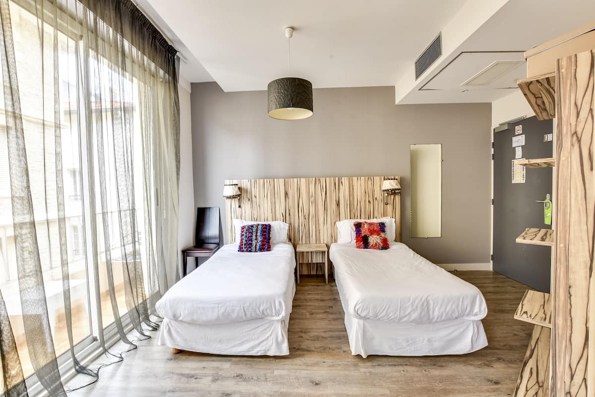 A modern hotel room with two single beds, a wooden headboard, a desk with a chair, and a large window with sheer curtains.