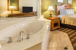 Una habitación de hotel con una bañera grande, una mesita de noche de madera con una lámpara, una cama con ropa blanca y un televisor sobre un mueble de madera.