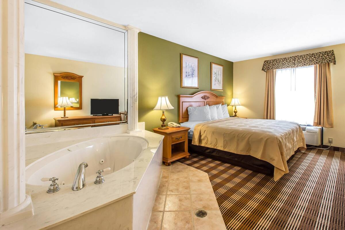 A hotel room with a large bathtub, a bed with a headboard, two bedside lamps, and a window with curtains.