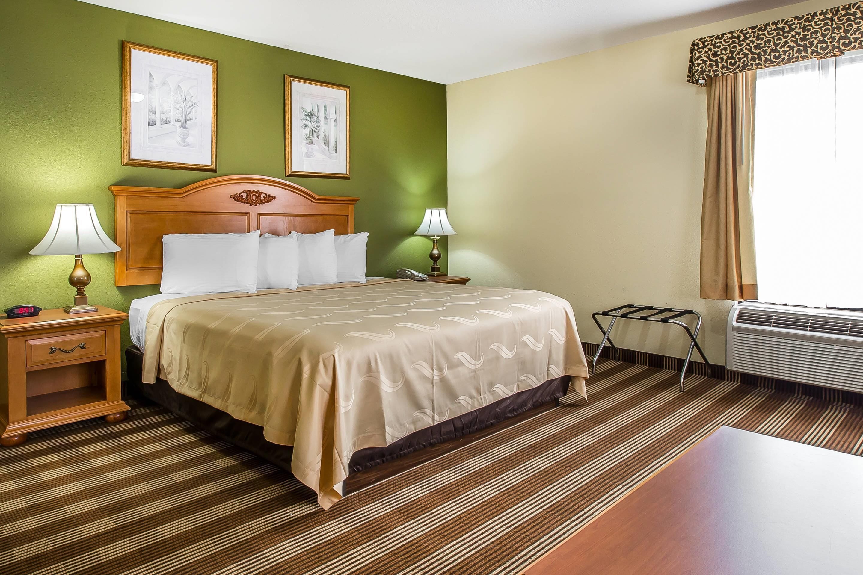 A hotel room with a bed, two bedside lamps, a nightstand, a window with curtains, and an air conditioning unit.