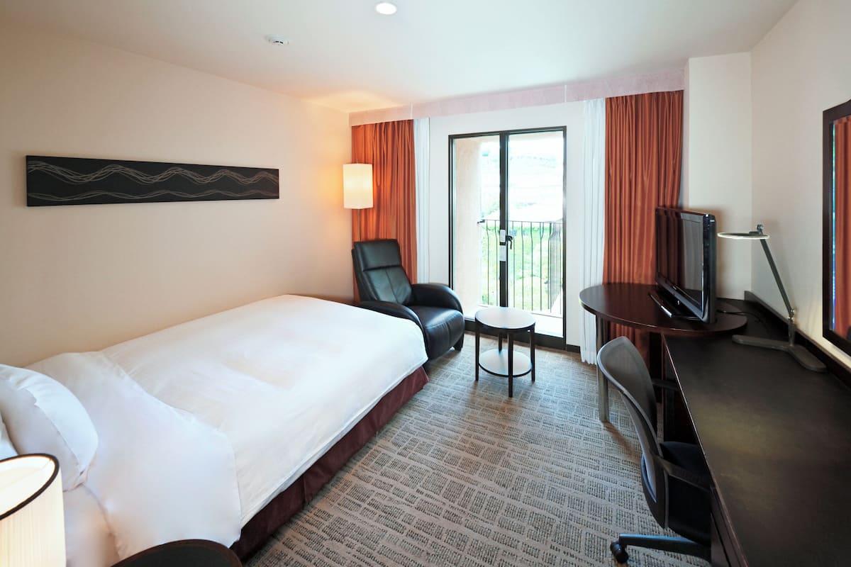 A hotel room with a bed, a desk with a TV, a chair, a small table, and a balcony with curtains.