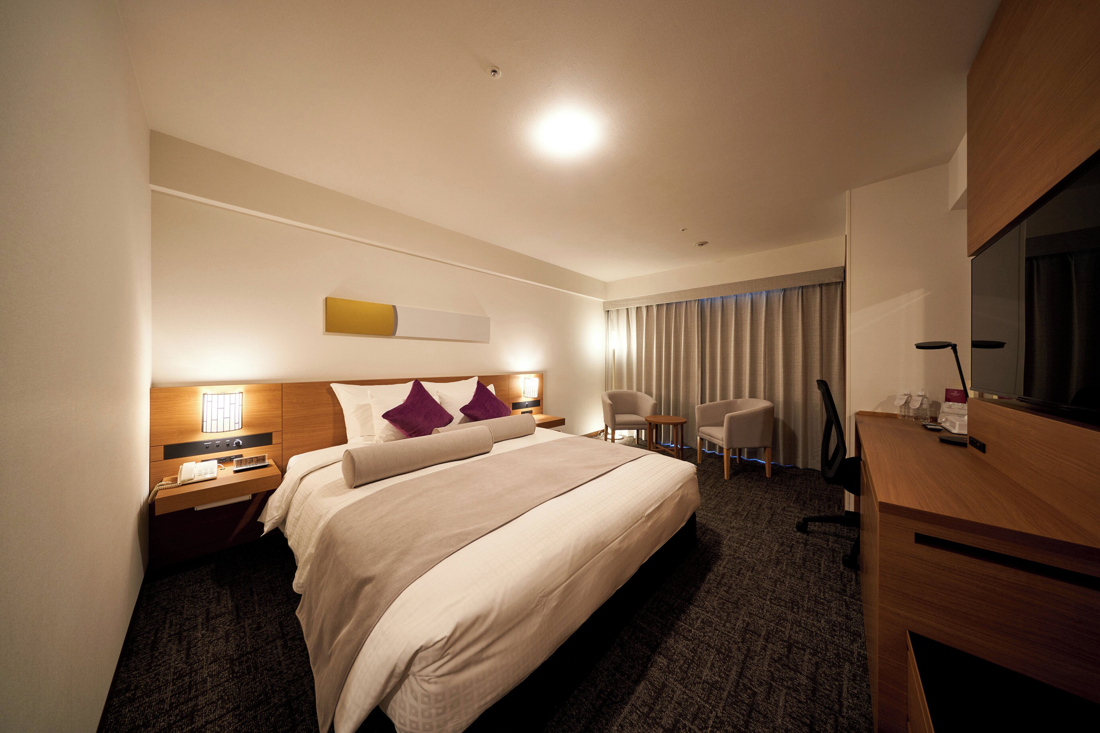 A hotel room with a bed, bedside table, lamp, chair, desk, and TV.