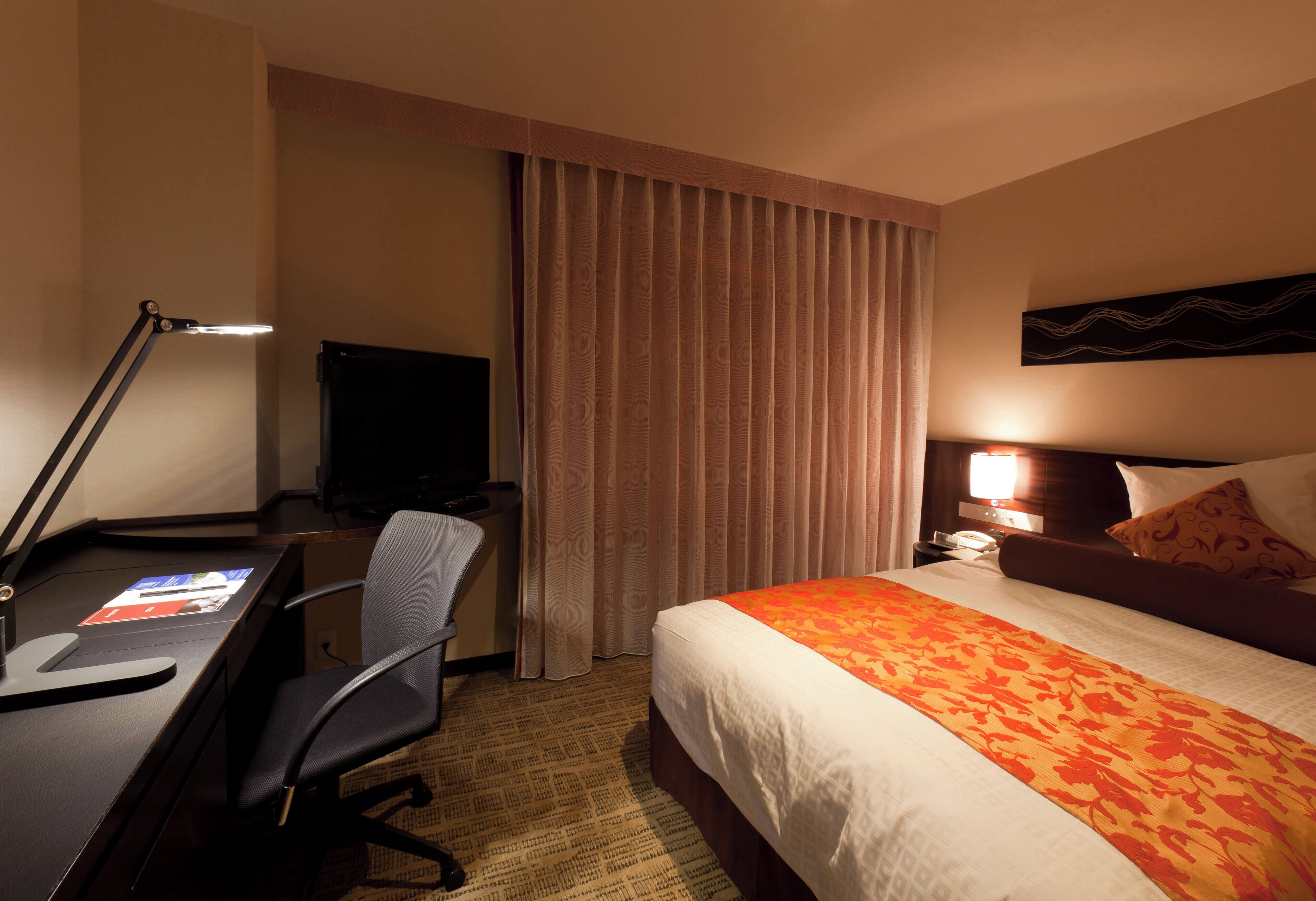 A hotel room with a desk, chair, bed, curtains, and a television.