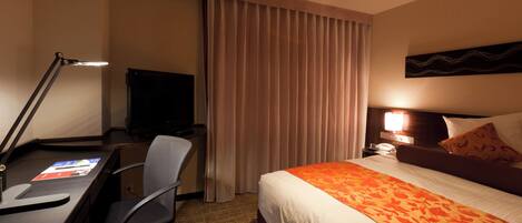 A hotel room with a desk, chair, bed, curtains, and a television.