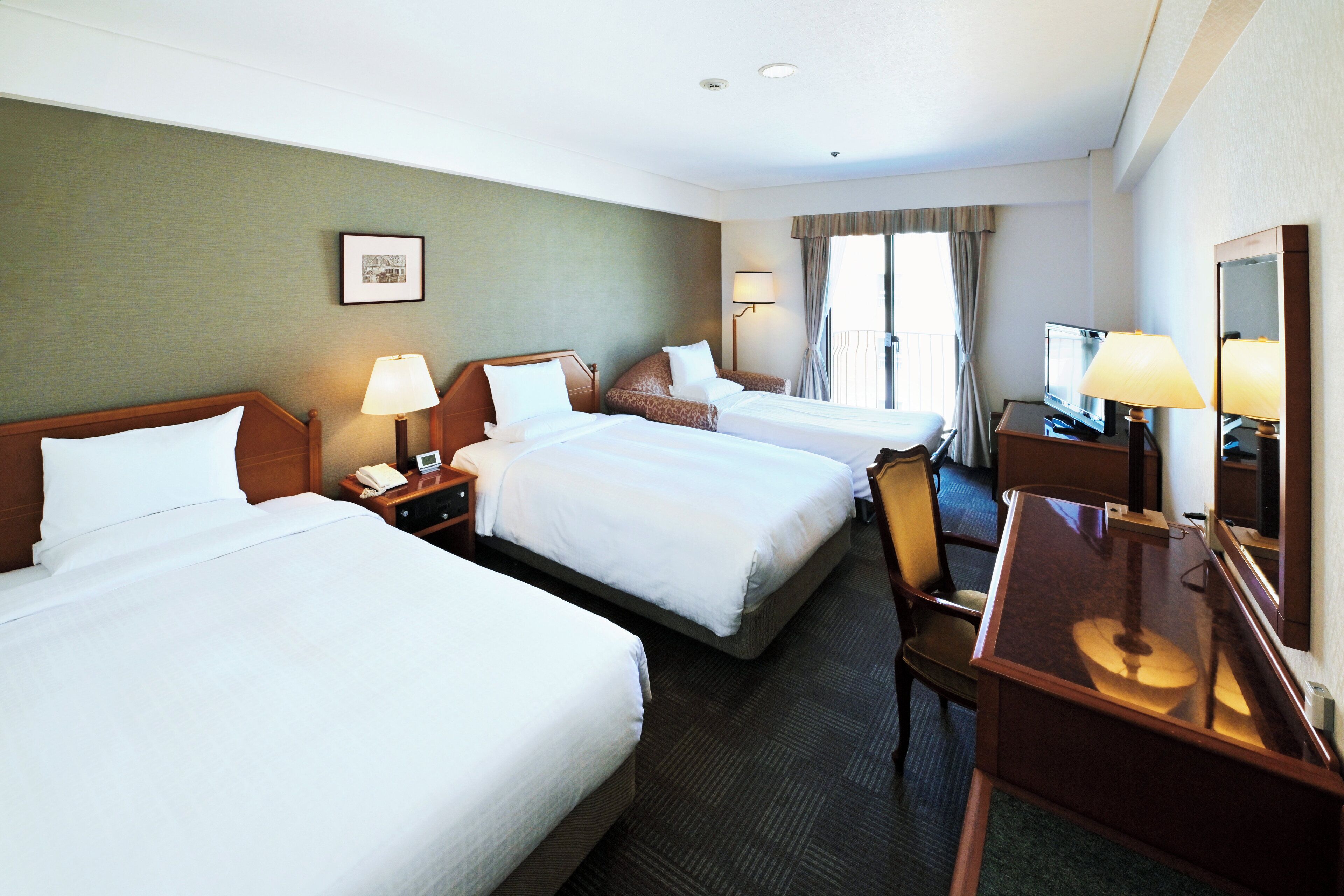 A hotel room with three beds, a desk, a chair, a TV, and a window with curtains.