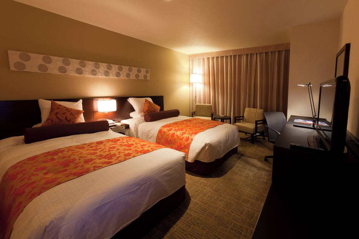 A hotel room with two beds, a desk, a chair, a TV, and a lamp.