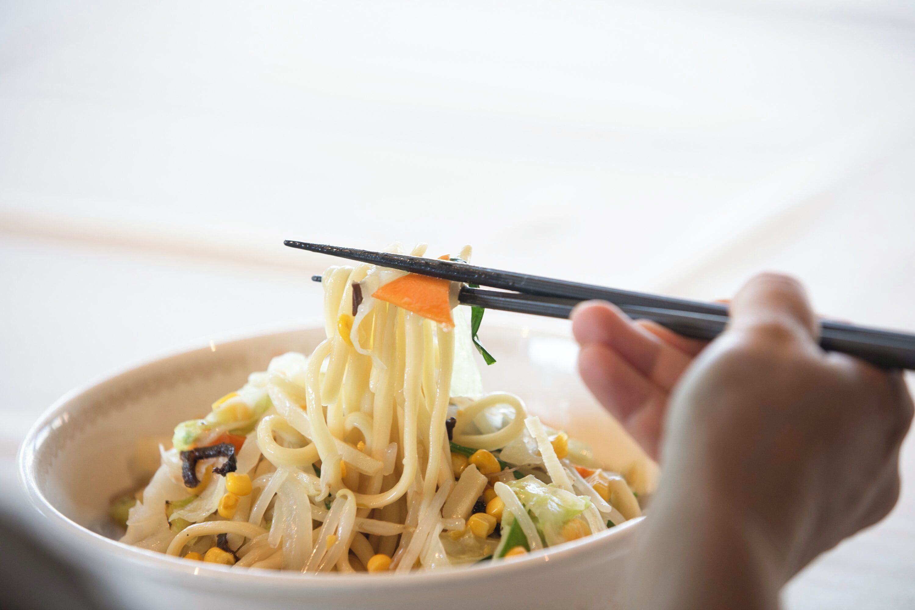 A bowl of noodles with vegetables and corn being lifted with chopsticks.