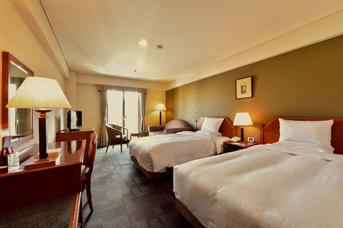 A hotel room with two beds, a desk, a chair, a TV, and a window with curtains.