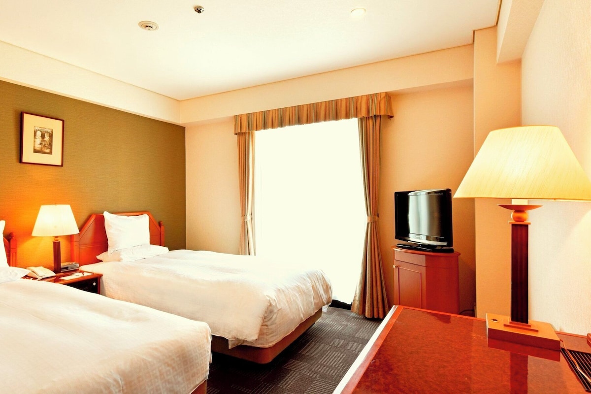 Habitación de hotel con dos camas, un televisor sobre un mueble bajo y un escritorio con lámpara.