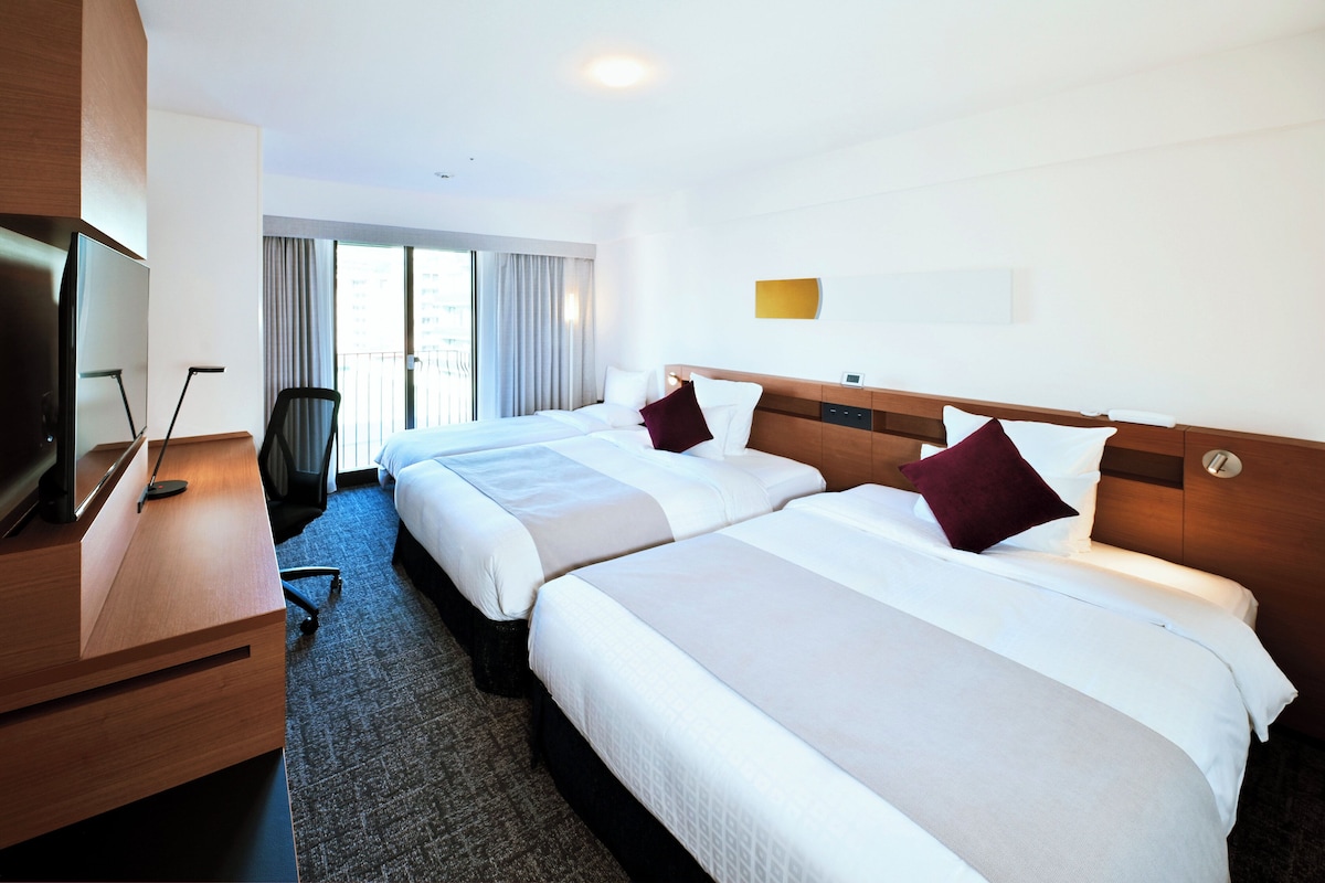 Habitación de hotel con dos camas, un escritorio con televisor y vistas a la ciudad a través de amplias ventanas.