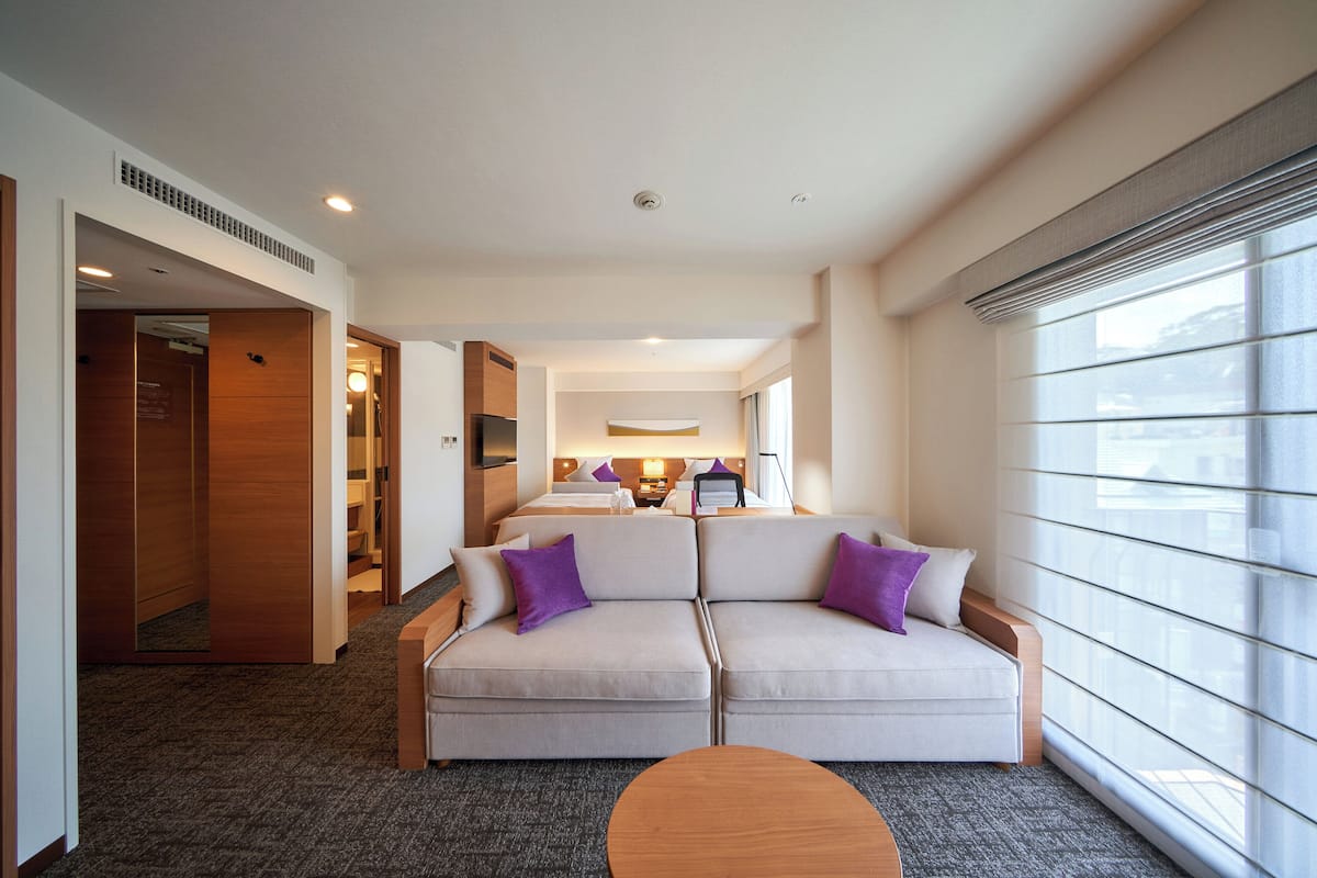A hotel room with a sofa, a small round table, a bed, and a window with blinds.