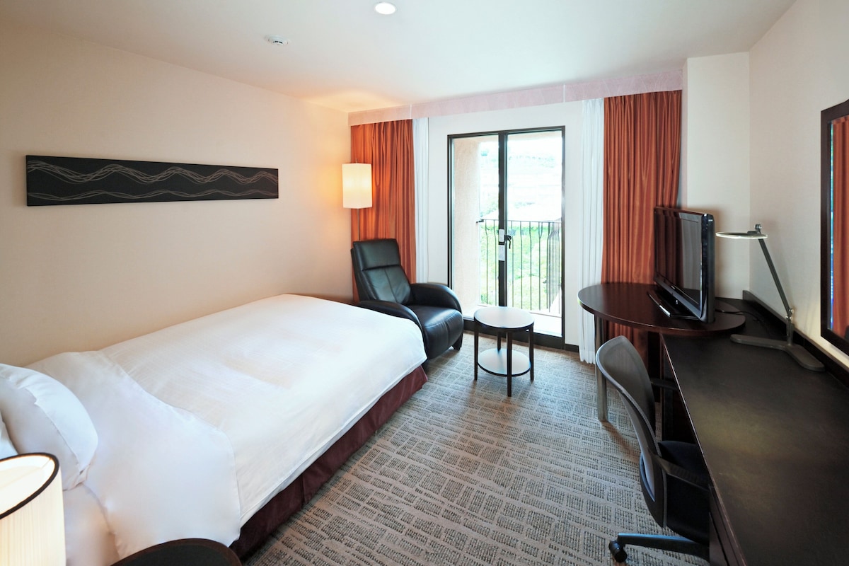 Habitación de hotel con cama, escritorio con televisor, silla, mesita y balcón con cortinas.