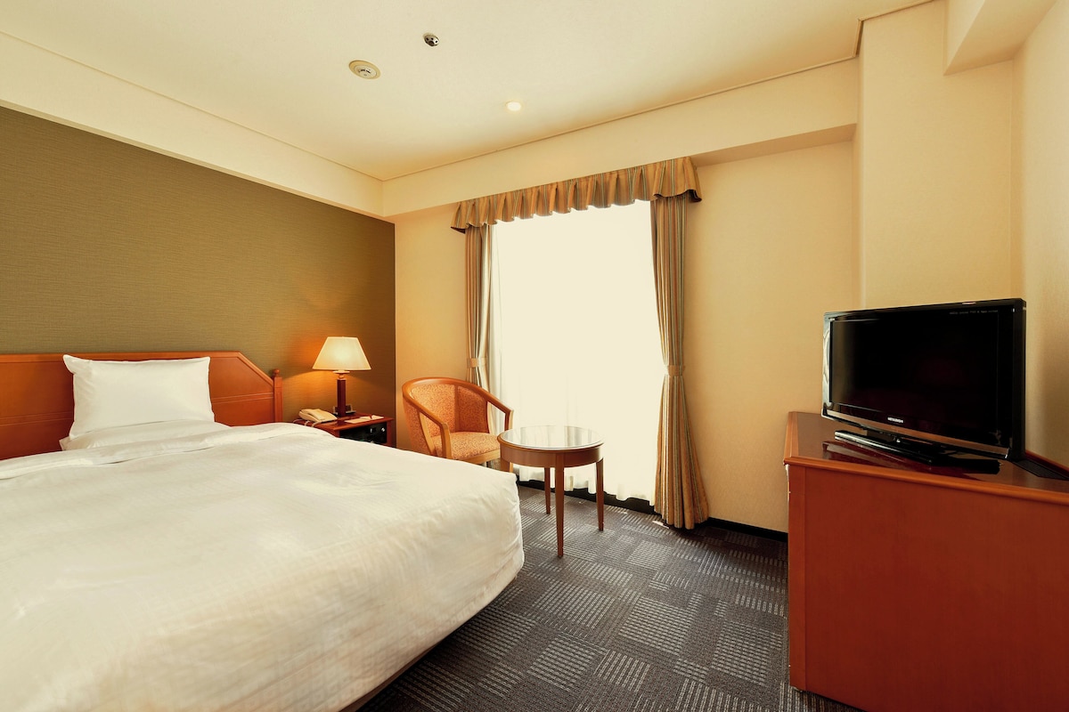Habitación de hotel con una cama grande, un televisor sobre un soporte de madera, una mesita, una silla y una mesita de noche con lámpara.