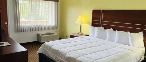 A hotel room with a large bed, a bedside table with a lamp, a window with blinds, and a wall-mounted heater.