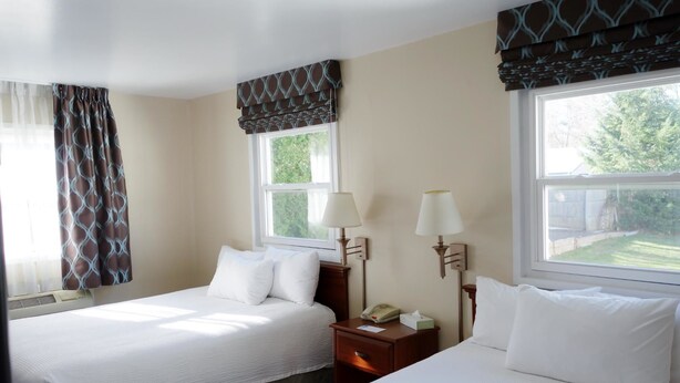 A hotel room with two beds, a bedside table with a lamp, and a window with a view of greenery. - Monument Mountain Motel (Great Barrington)