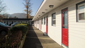 A row of motel rooms with red doors, a covered walkway, and a view of mountains in the background. - Monument Mountain Motel (Great Barrington)