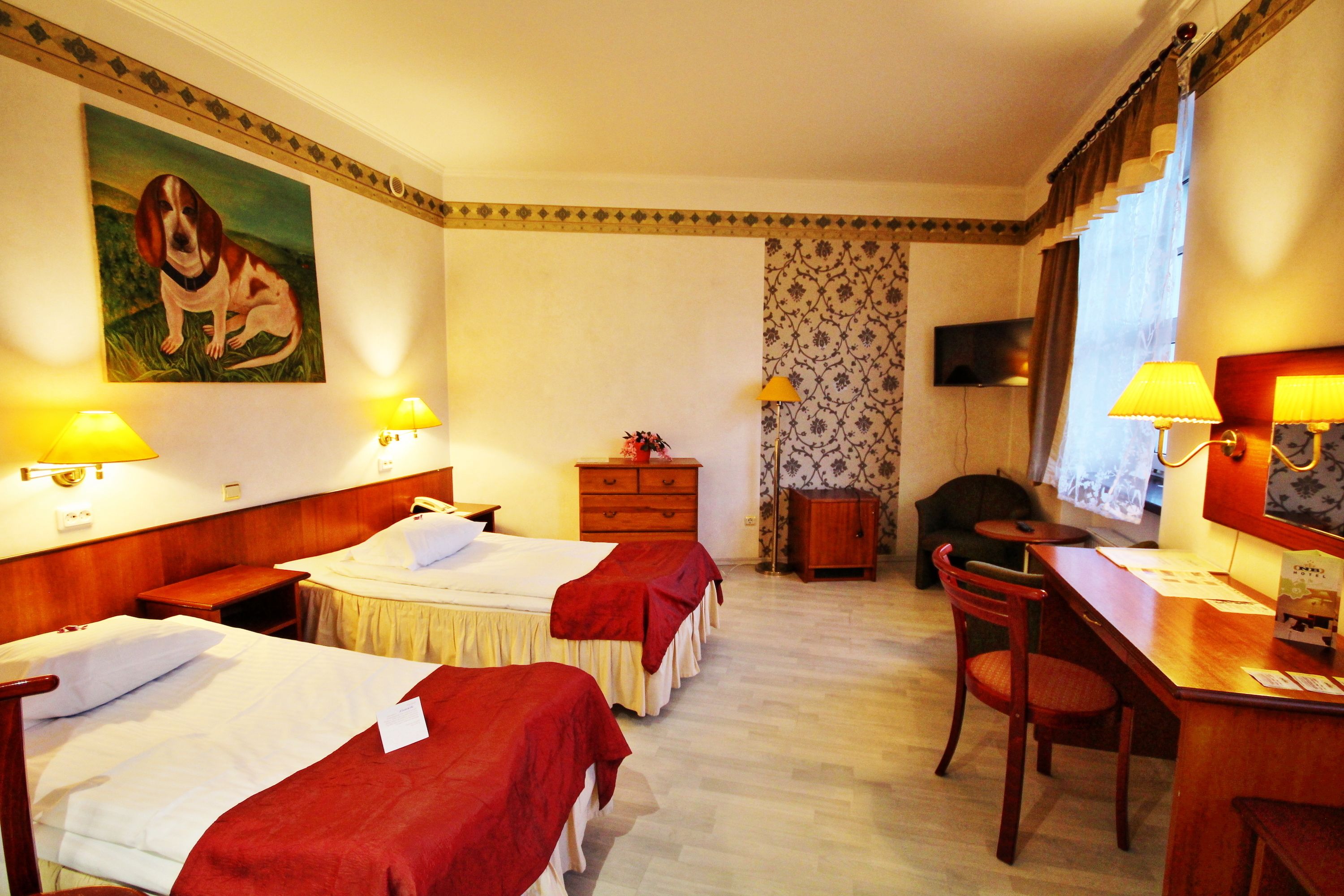 A hotel room with two beds, a desk, a chair, a dresser, and a painting on the wall.