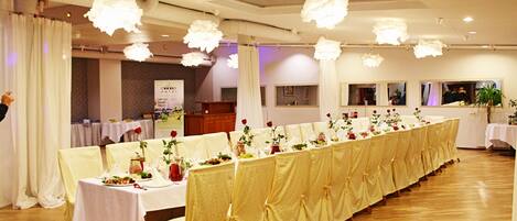 A banquet hall with long tables covered in white tablecloths, adorned with floral centerpieces and red roses.