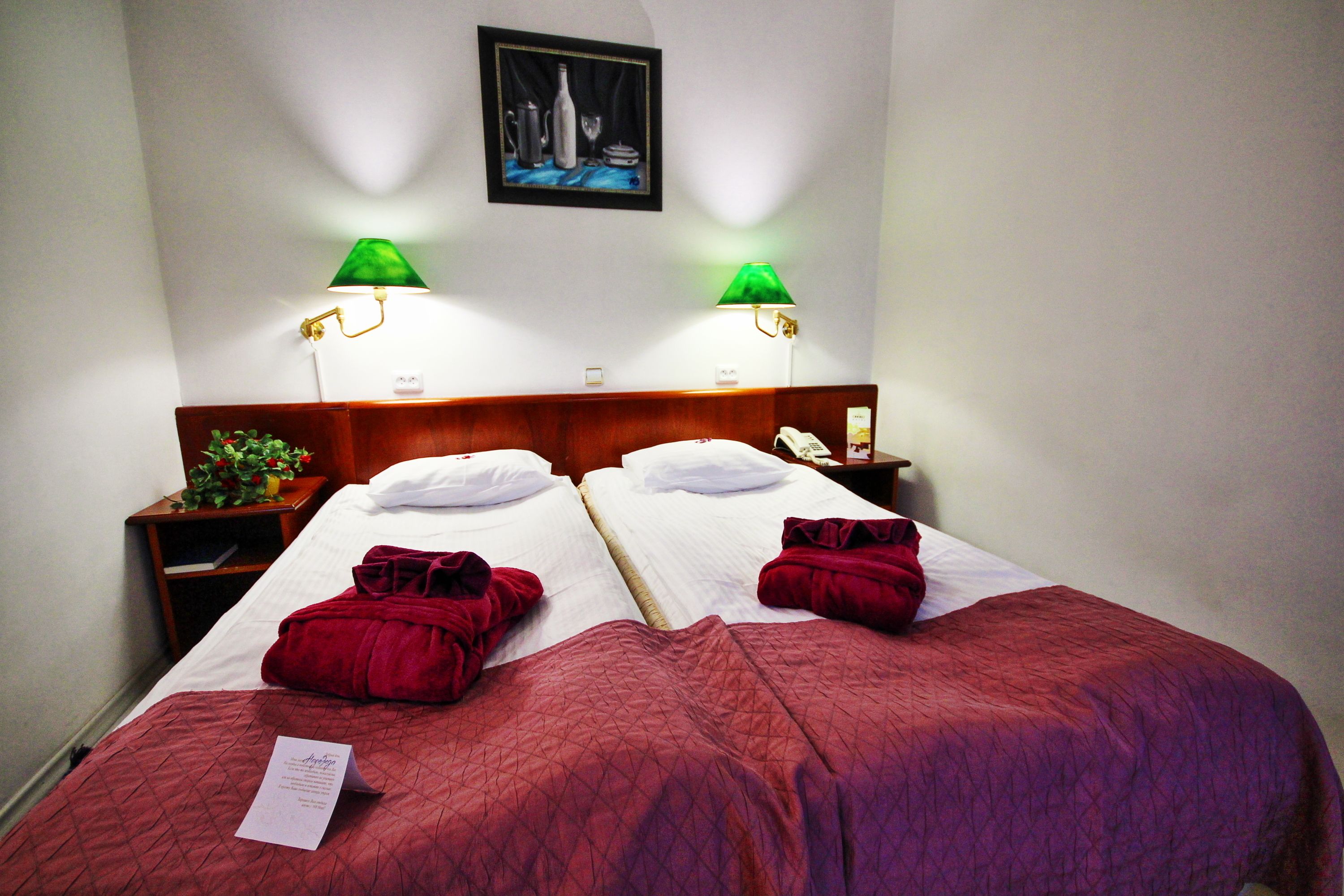 Habitación de hotel con dos camas, cada una con una colcha y almohadas rojas, una mesita de noche con lámpara y un cuadro enmarcado en la pared.