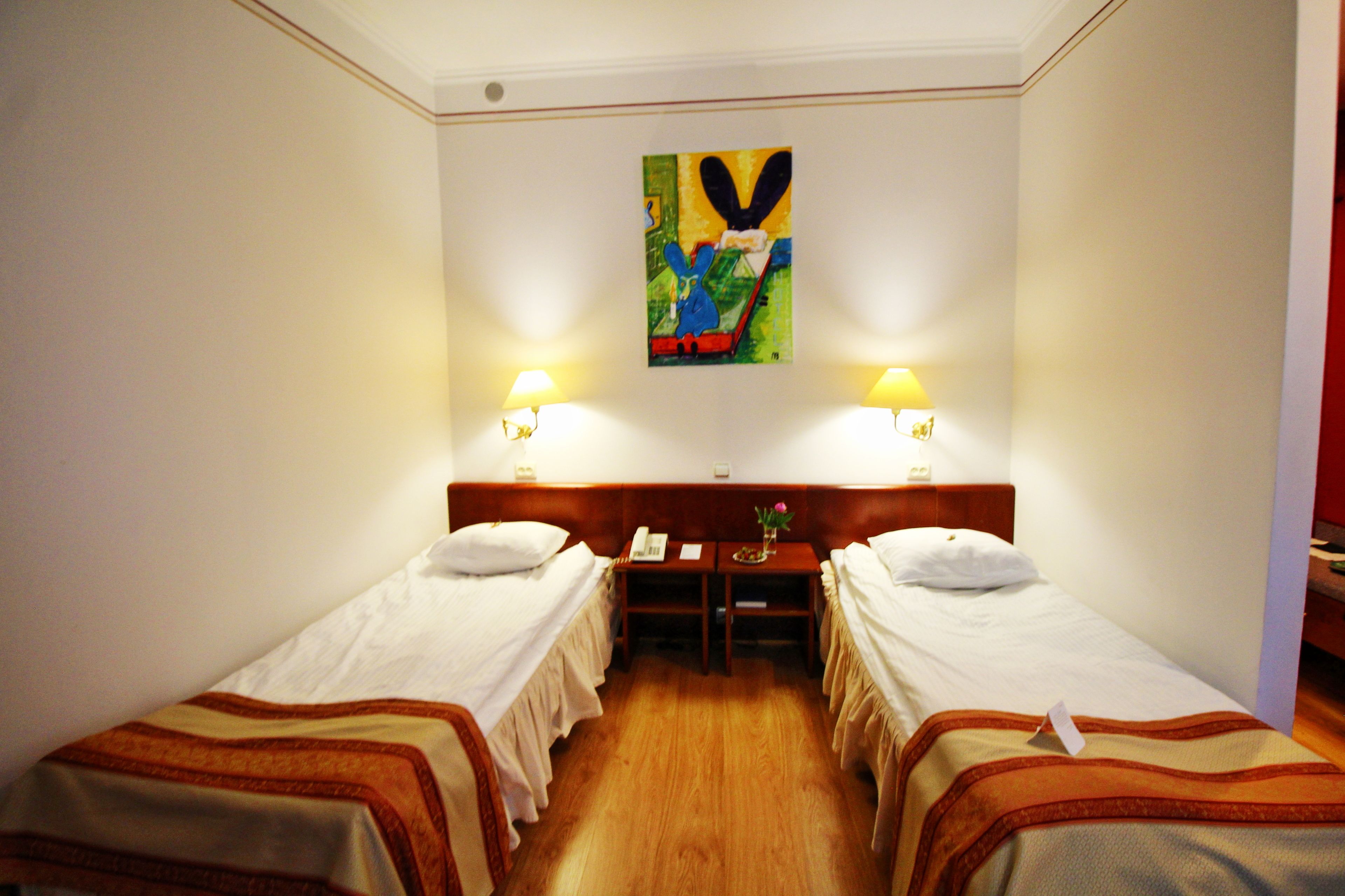 A hotel room with two single beds, a wooden desk, two bedside lamps, and a colorful abstract painting on the wall.