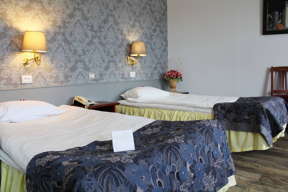A hotel room with two beds, a nightstand, a lamp, a vase with flowers, and a framed picture on the wall.