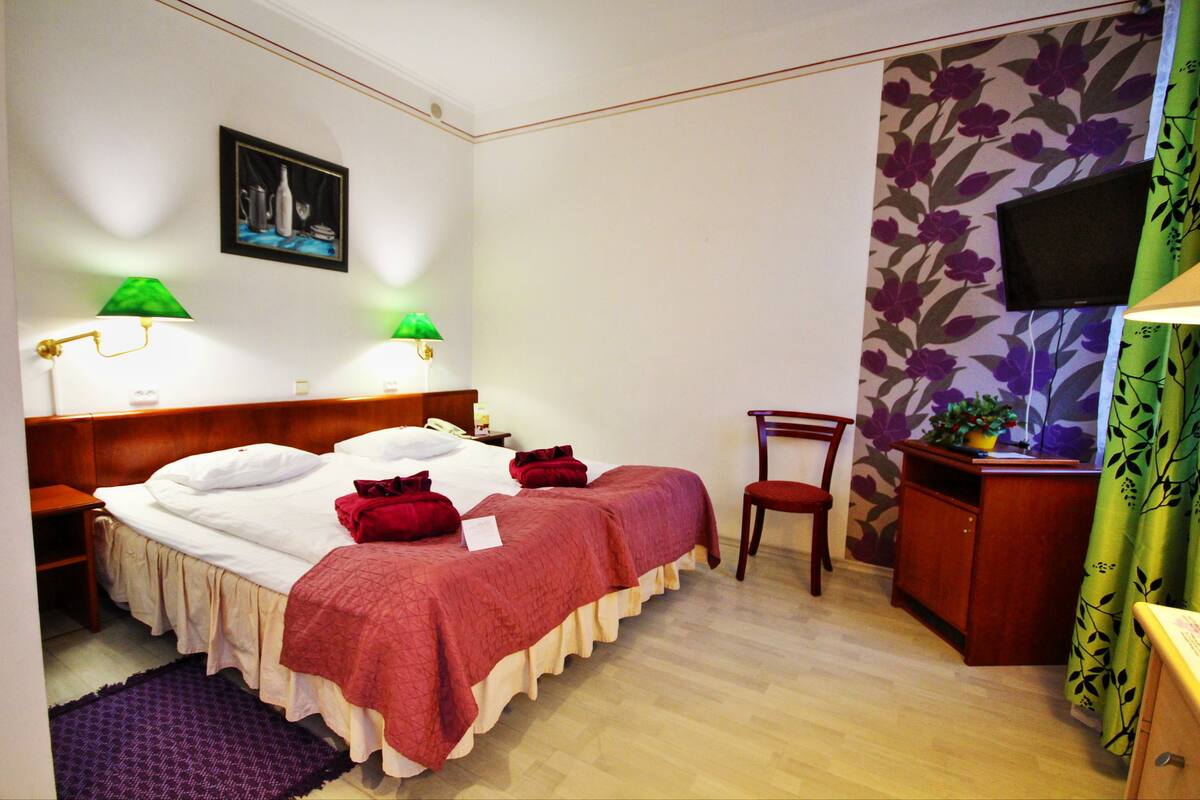 A hotel room with a bed, two bedside lamps, a chair, a desk, a television, and a floral wallpaper.