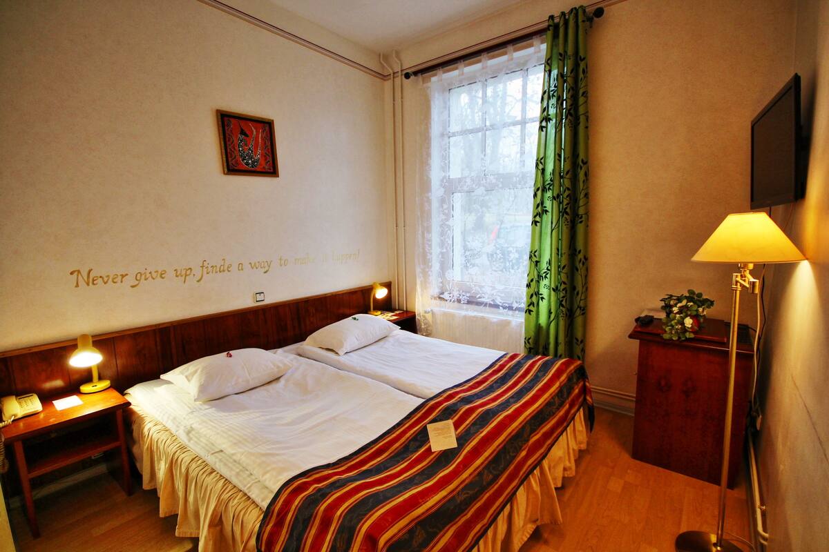 A hotel room with a double bed, a bedside table with a lamp, a small desk with a telephone, a window with green curtains, and a motivational quote on the wall.