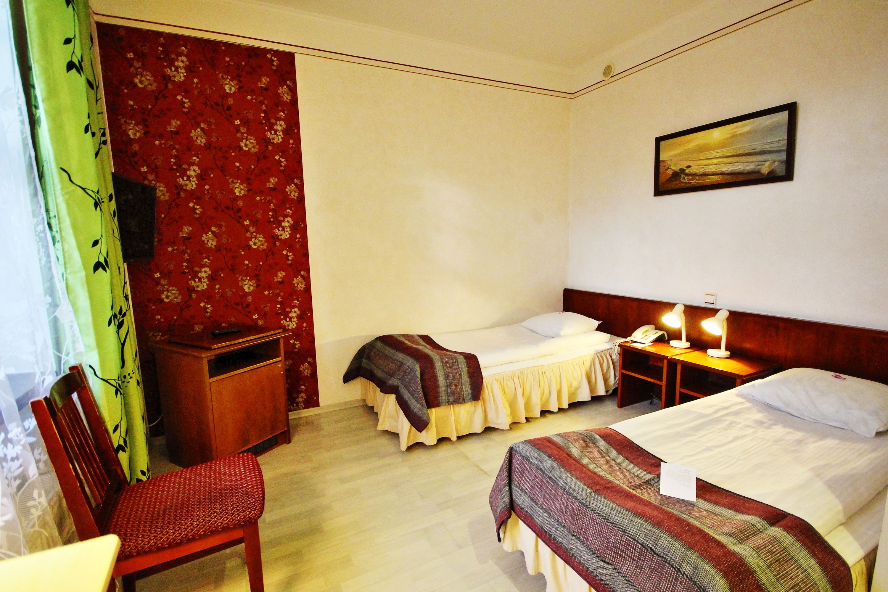 A hotel room with two single beds, a wooden nightstand with a lamp, a desk with a telephone, a chair, and a painting on the wall.