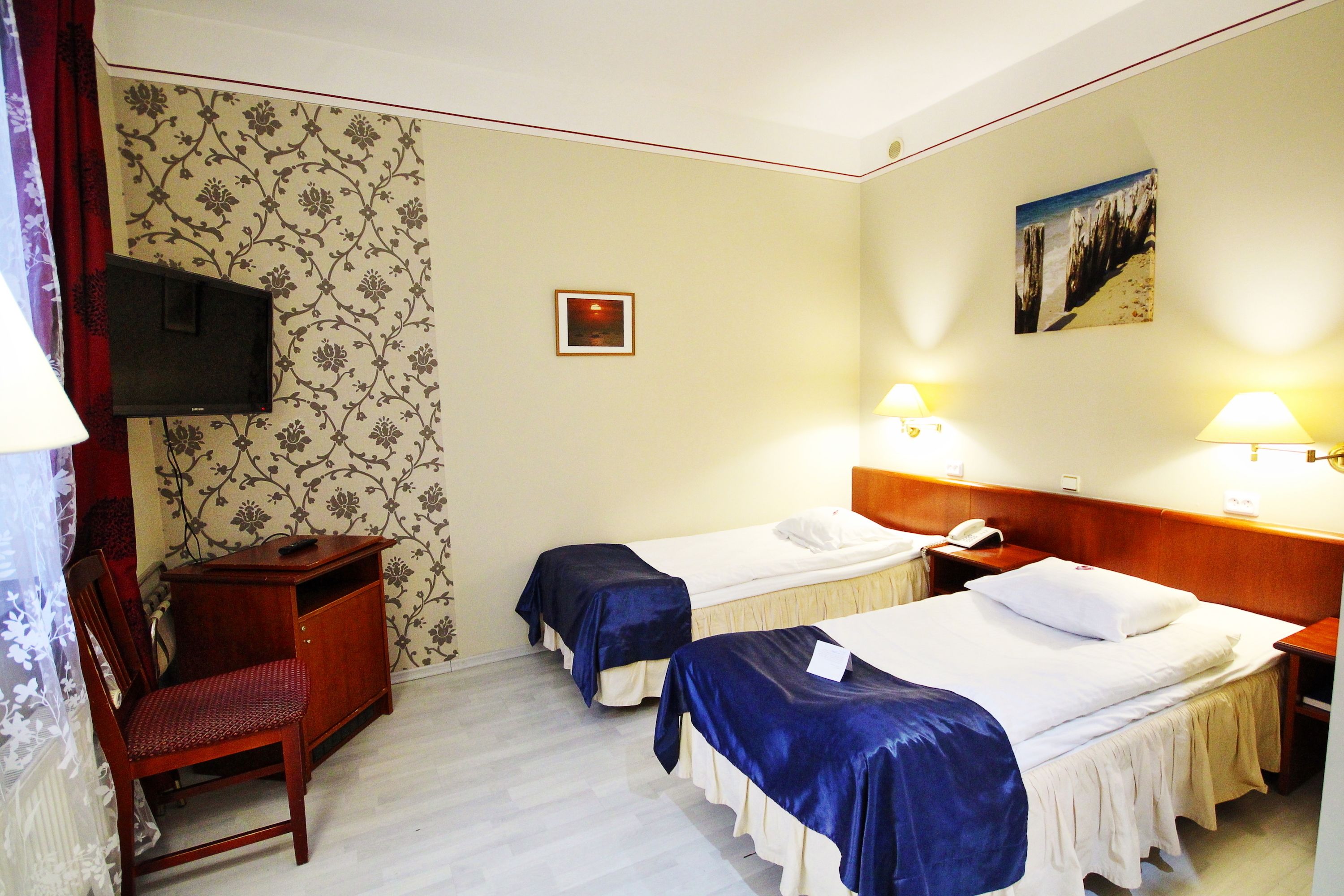 A hotel room with two beds, a desk, a chair, a TV, and two lamps.