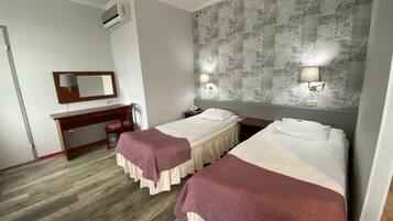 Hotellihuone, jossa on kaksi sänkyä, työpöytä, tuoli, peili ja seinälle asennettu ilmastointilaite.