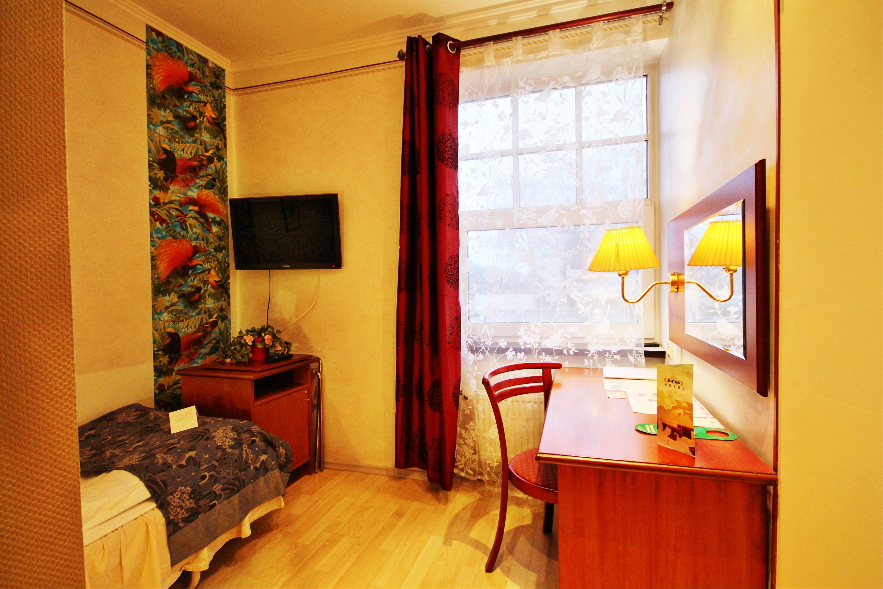 Une petite pièce comprenant un lit, un bureau, une chaise, un téléviseur et une fenêtre avec des rideaux.