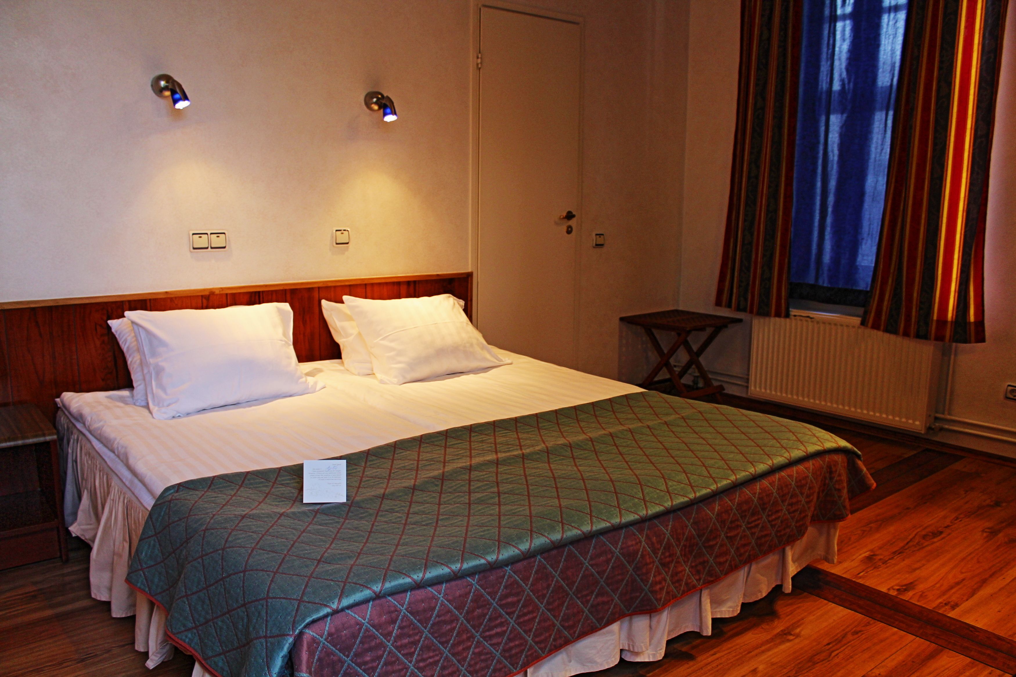 Una habitación de hotel con cama, mesita de noche, ventana con cortinas e iluminación en la pared.