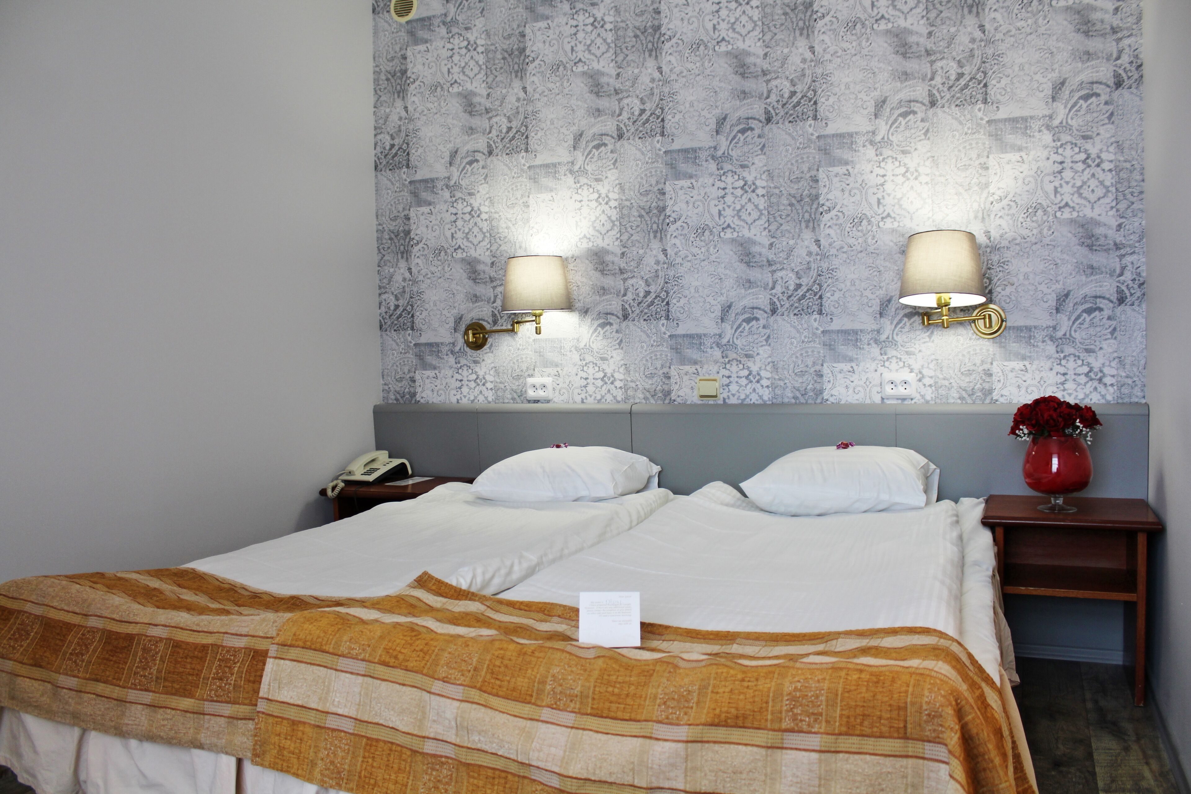 A hotel room with two beds, a nightstand with a lamp, a vase with flowers, and patterned wallpaper.