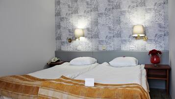 A hotel room with two beds, a nightstand with a lamp, a vase with flowers, and patterned wallpaper.
