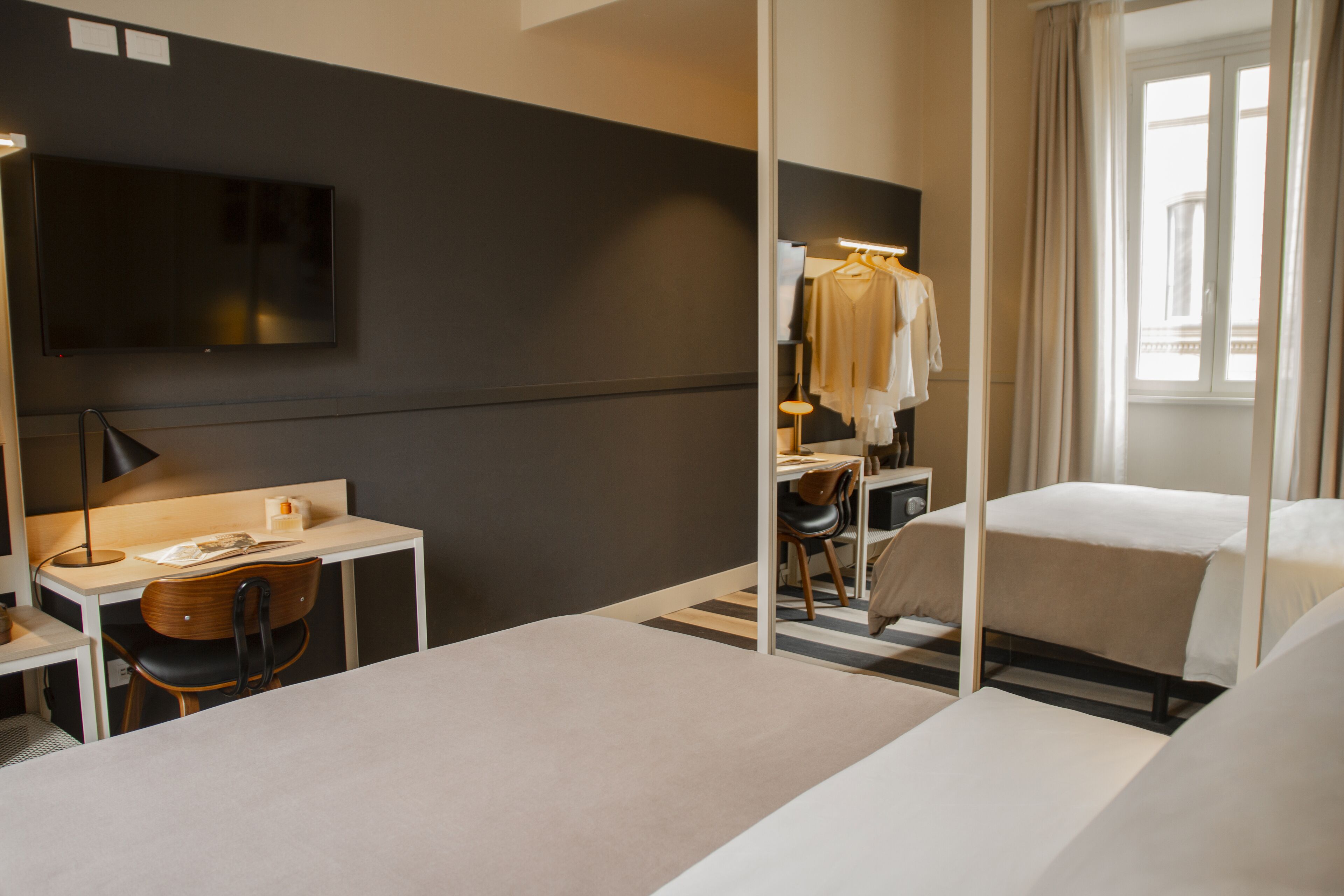 A hotel room with a bed, a desk with a chair, a TV mounted on the wall, and a window with curtains.