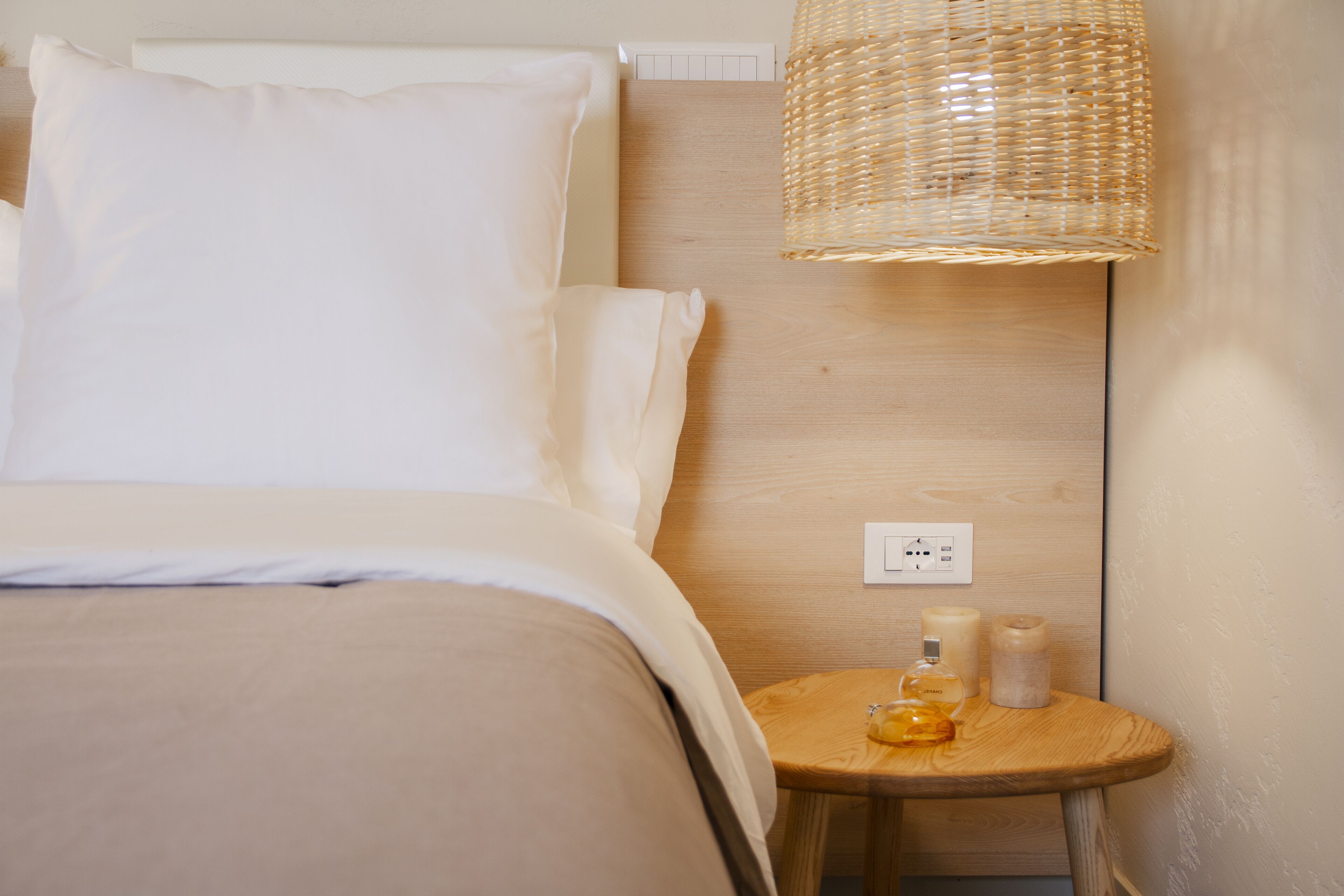 A neatly made bed with white linens, a bedside table with a bottle and a candle, and a woven lamp.