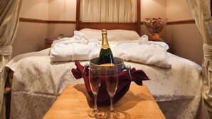A neatly made bed with a bottle of champagne and two glasses on a wooden table, a vase of flowers, and a bedside table with a lamp. - The Old Mill Hotel (Bury)