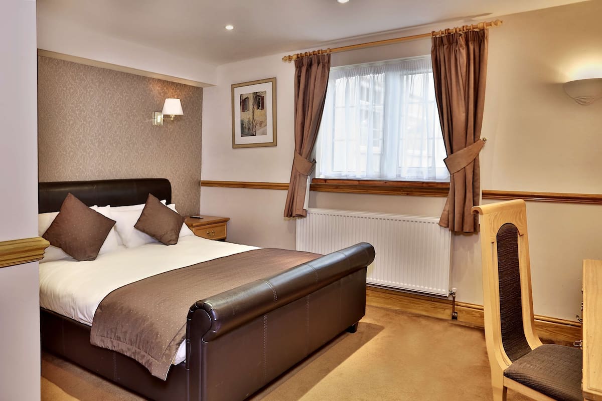 A neatly arranged hotel room with a large bed, a bedside table, a chair, a window with curtains, and a wall-mounted lamp.