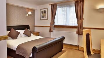 Habitación de hotel ordenada con una cama grande, una mesita de noche, una silla, una ventana con cortinas y una lámpara de pared.
