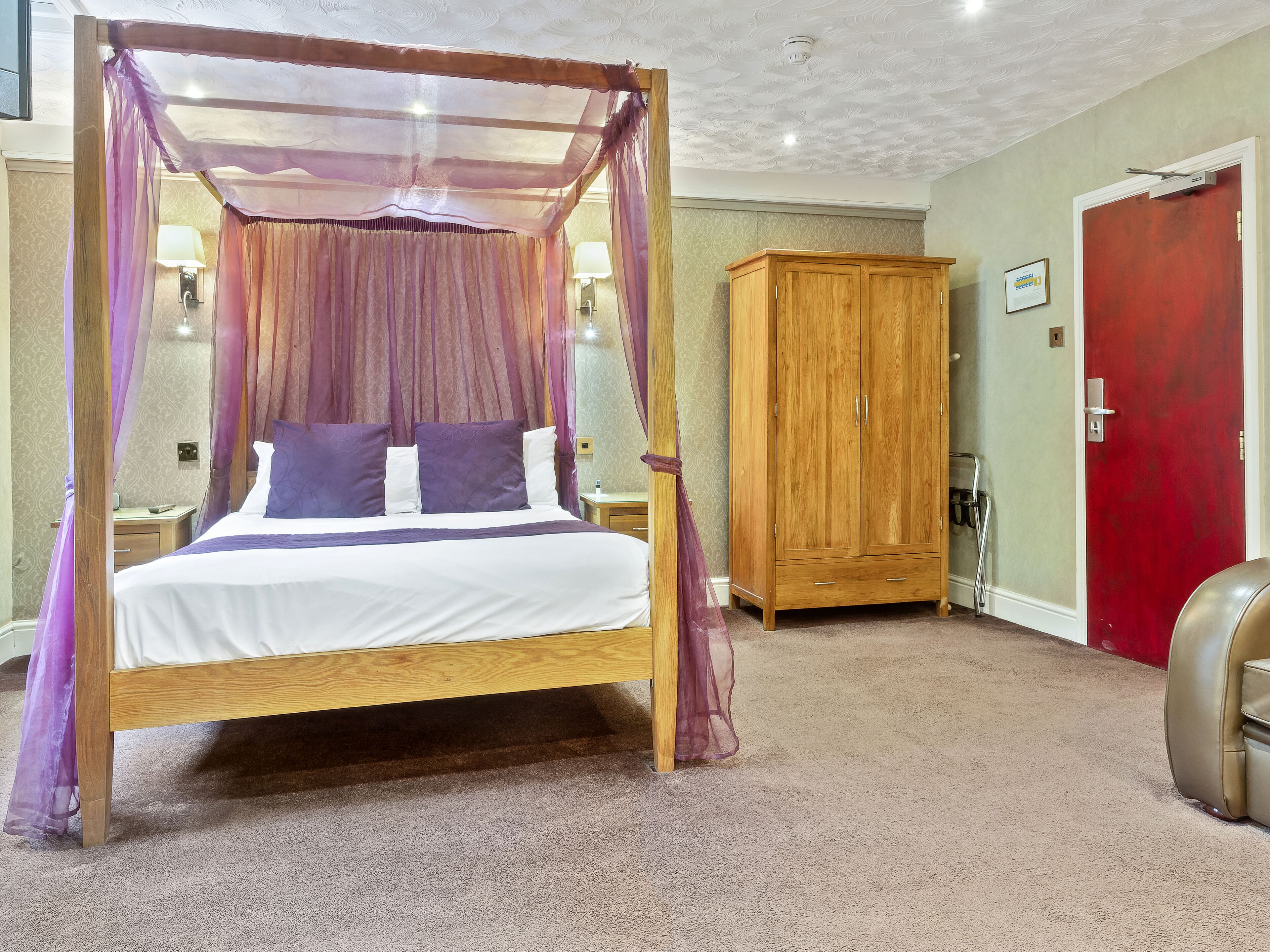 A four-poster canopy bed with a wooden frame and sheer curtains, a wooden wardrobe, and a red door in a hotel room.