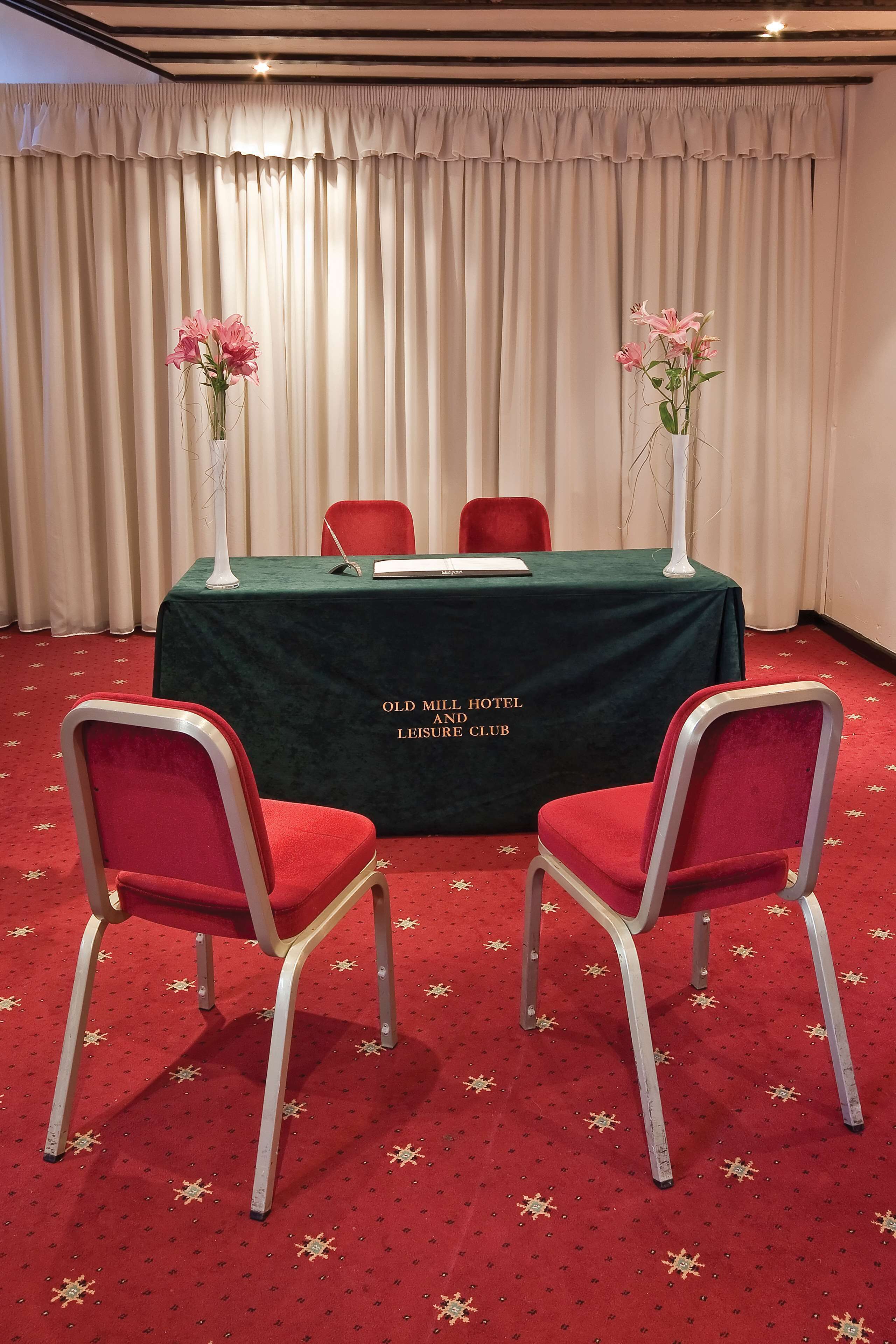 Une salle de conférence avec une table recouverte d’une nappe verte, des chaises rouges et deux vases ornés de fleurs roses.