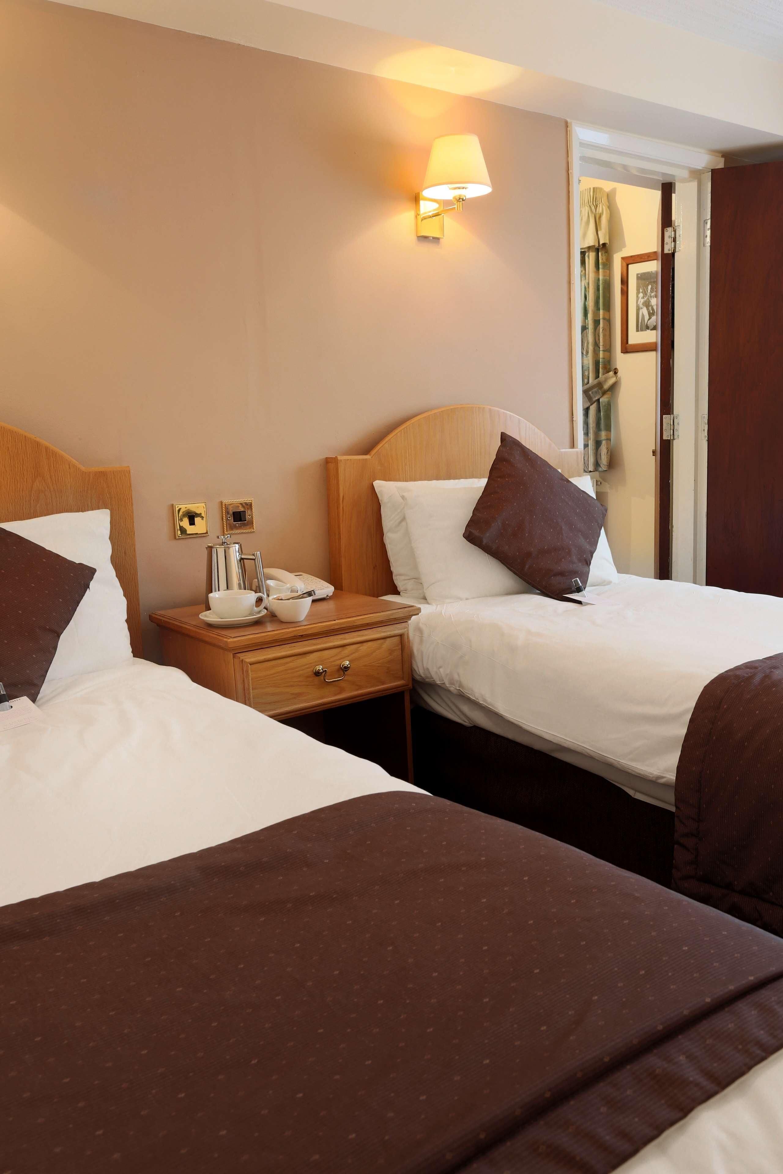 A hotel room with two beds, a bedside table with a lamp, a telephone, and a small table with a cup and saucer.
