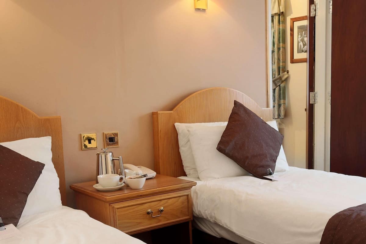 A hotel room with two beds, a bedside table with a lamp, a telephone, and a small table with a cup and saucer.