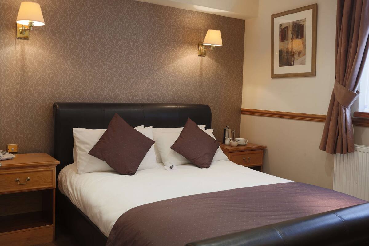 A hotel room with a large bed, two bedside tables, a nightstand, a framed picture on the wall, and a window with curtains.