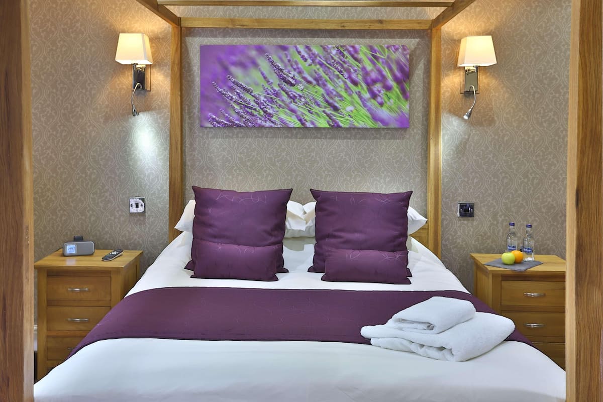 A neatly made bed with purple pillows, a wall-mounted artwork, bedside lamps, a nightstand with a clock, and a small table with fruit and bottles.